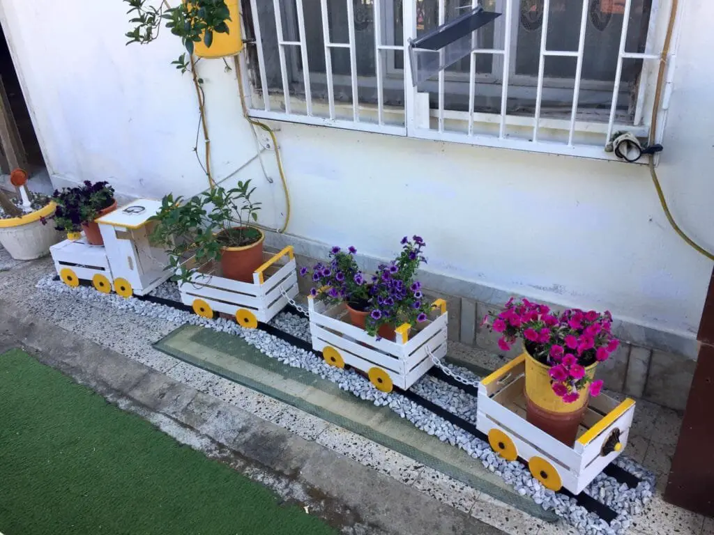 Coltivare l’Orto sul Balcone: Idee, Consigli e Piante Giuste 9 Cassette di legno colorate riutilizzate come fioriere mobili, posizionate lungo un muro esterno per creare un orto decorativo e funzionale.