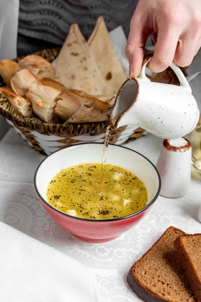 Salmoriglio Siciliano: Ricetta, Usi e Ingredienti Freschi 2 Scena di casa: sto versando del salmoriglio profumato in una ciotola per intingere il pane fatto in casa, con una cesta di pane rustico e pane carasau a portata di mano. Mi piace servire il salmoriglio così quando arrivano amici, per una cena senza formalità ma piena di sapori veri.
