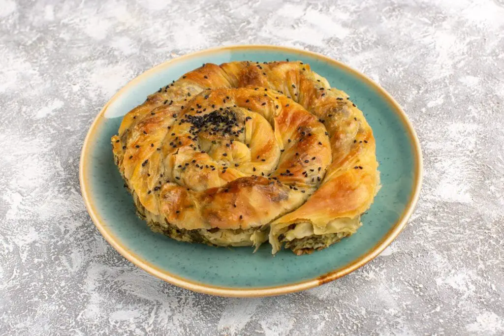 Borek Turco di Verdure: Ricetta Facile, Varianti e Consigli 1 Borek turco di verdure arrotolato, servito su piatto turchese, crosta dorata e semi di sesamo, ripieno visibile all’estremità.