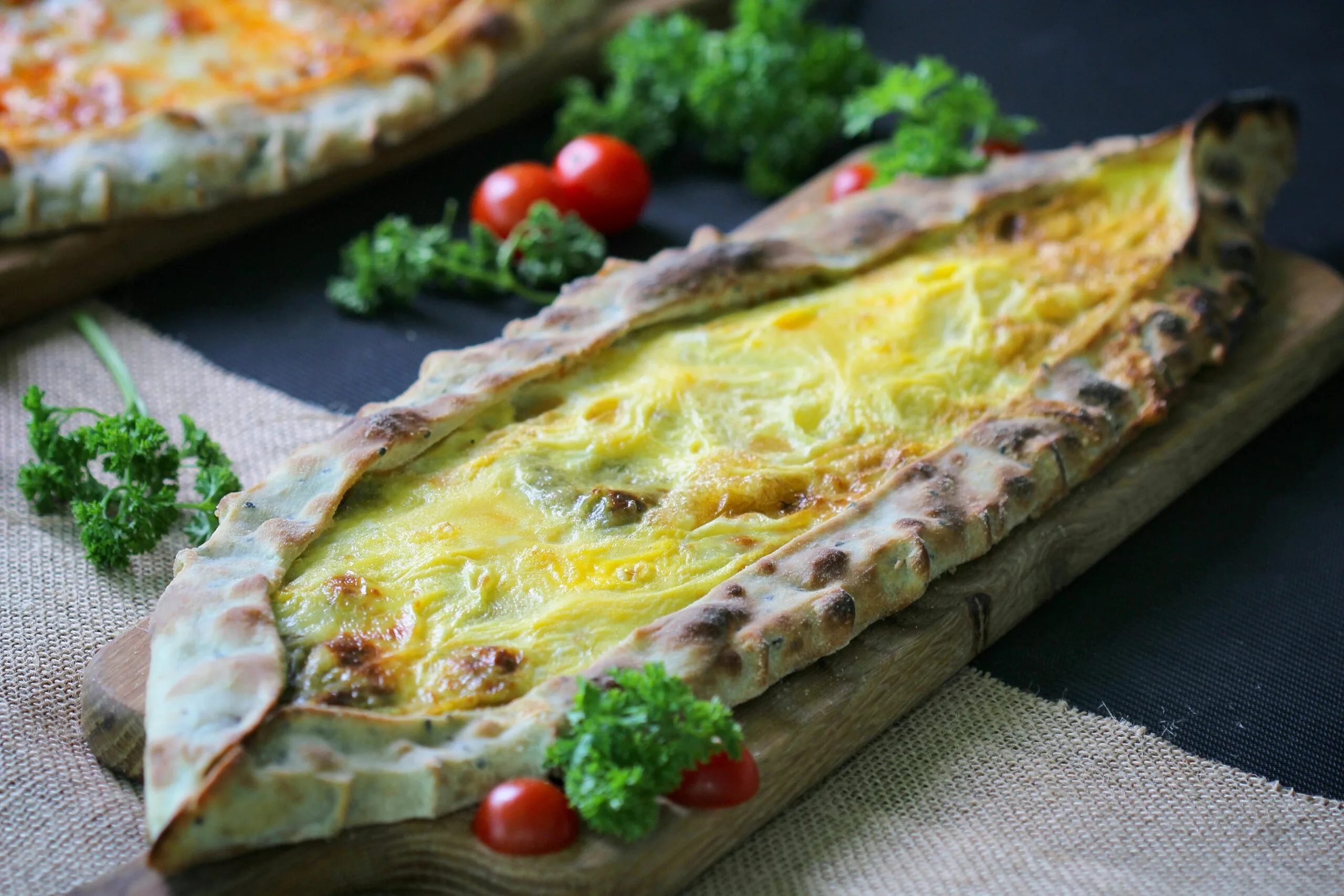 Pide al formaggio appena sfornato, con crosta dorata e ripieno di formaggio fuso, servito su un tagliere con pomodorini e prezzemolo fresco.