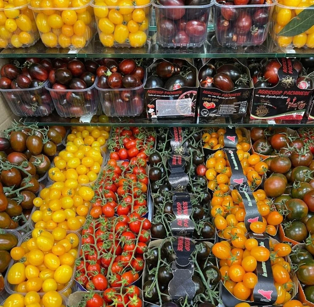 10 Segreti per Coltivare Pomodorini Biologici in Orto e Vaso 2 Espositore in un negozio con diverse confezioni di pomodorini ciliegino di varie colorazioni, tra cui gialli, rossi, arancioni e neri, disposti su scaffali in plastica trasparente.
