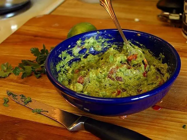 Ricetta Guacamole Messicano con Avocado e Ortaggi di Casa 3 Guacamole fatto in casa in ciotola blu con pezzi di pomodoro e lime sullo sfondo
