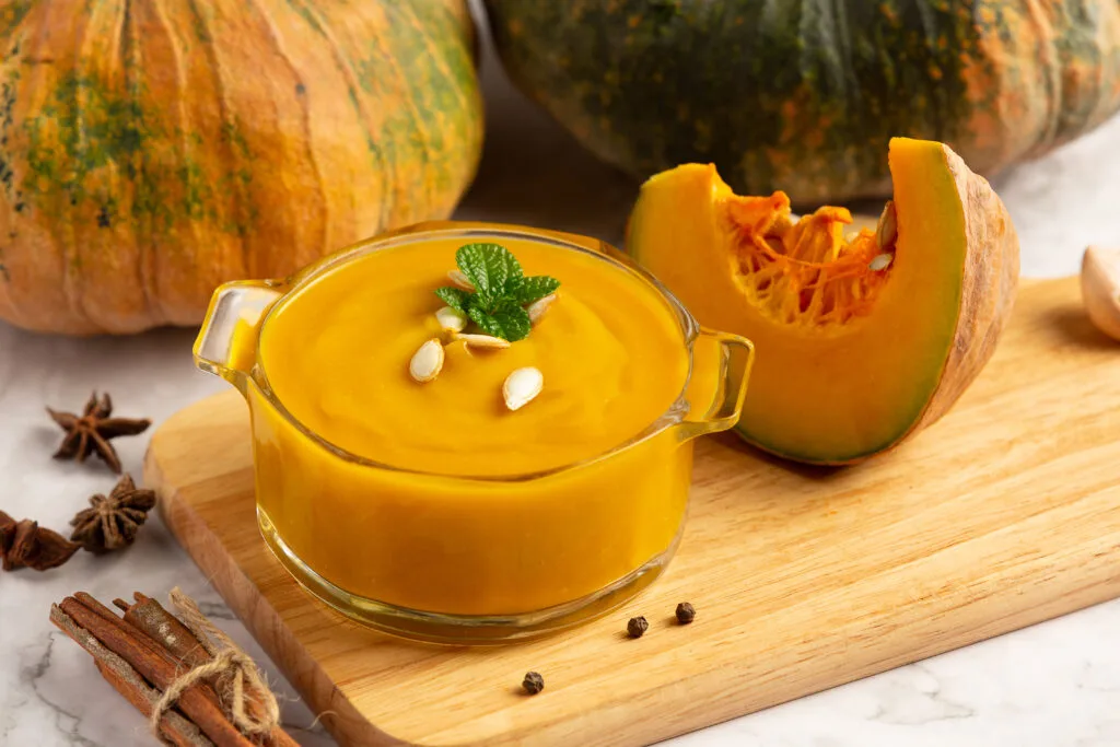 Corba di zucca turca: ingredienti, ricetta e coltivazione 7 Crema di zucca in ciotola di vetro, decorata con pinoli e menta, zucche fresche sullo sfondo.