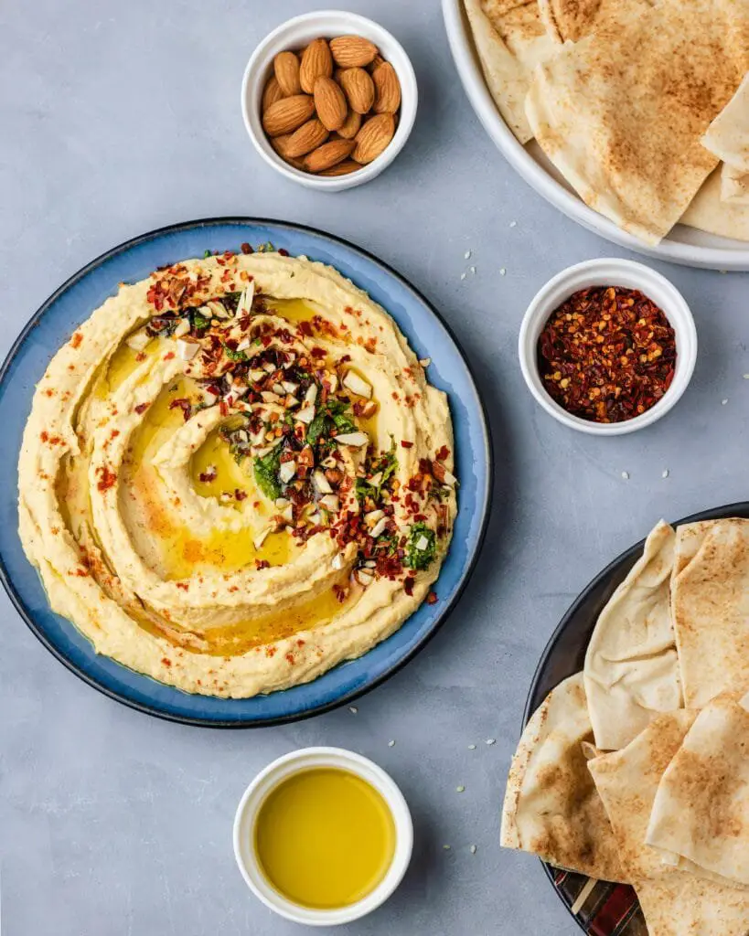 Hummus di Ceci Fatto in Casa: Ricetta Facile e Cremosa 3 Piatto di hummus di ceci guarnito con olio extravergine, frutta secca tritata e spezie, servito con pane pita su un tavolo chiaro